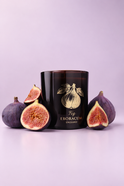 A dark amber glass candle jar with a gold fig illustration and the text 'Fig EBORACUM ENGLAND' on the front, styled with whole and halved purple figs on both sides against a soft lavender background.