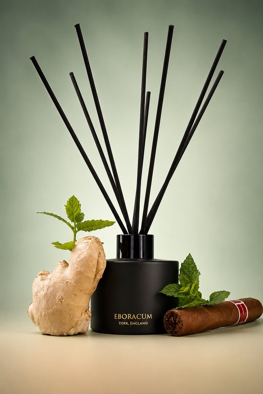 A black reed diffuser bottle with tall black reeds, styled with a ginger root, mint leaves, and a cigar on a beige surface against a soft green gradient background. The bottle is labeled 'EBORACUM' and 'MADE IN ENGLAND'.