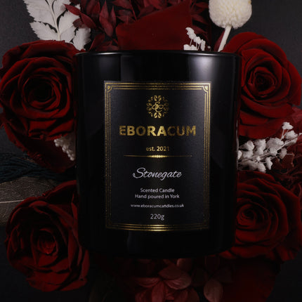 Black candle with gold label surrounded by red roses and decorative elements on a dark background
