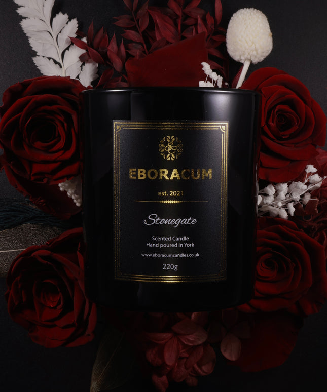 Black candle with gold label surrounded by red roses and decorative elements on a dark background