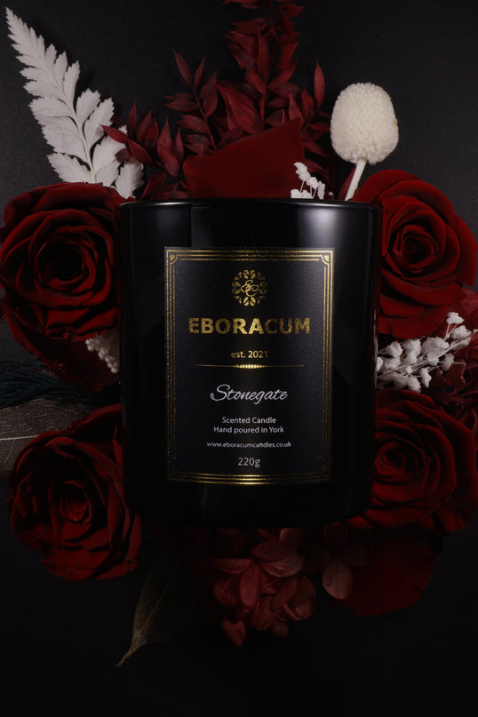 Black candle with gold label surrounded by red roses and decorative elements on a dark background