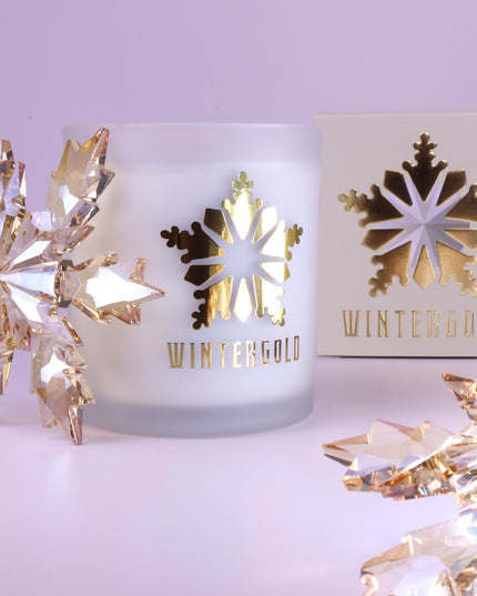 A frosted glass candle jar with a metallic gold snowflake cutout and the word 'WINTERGOLD' printed below, styled on a pale lavender surface with decorative crystal snowflakes and a matching box in the background.