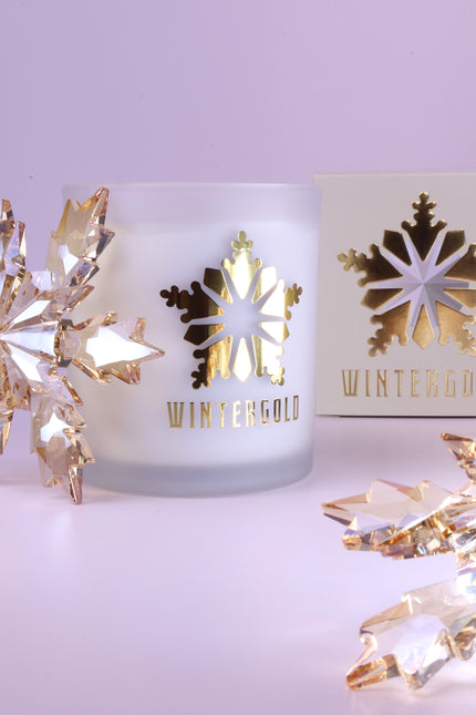A frosted glass candle jar with a metallic gold snowflake cutout and the word 'WINTERGOLD' printed below, styled on a pale lavender surface with decorative crystal snowflakes and a matching box in the background.