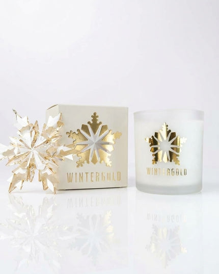A frosted glass candle jar with a gold foil snowflake and 'WINTERGOLD' text, shown next to its cream-colored packaging box and a decorative gold snowflake ornament on a reflective white surface.