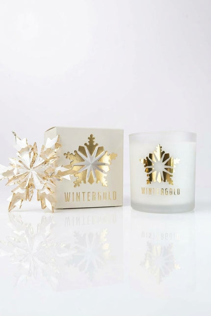 A frosted glass candle jar with a gold foil snowflake and 'WINTERGOLD' text, shown next to its cream-colored packaging box and a decorative gold snowflake ornament on a reflective white surface.