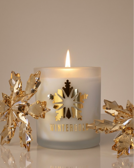 A frosted white candle with a lit flame, decorated with a gold snowflake and the word 'WINTERGOLD'. Gold crystal snowflake ornaments are arranged on both sides on a reflective surface against a warm beige background.